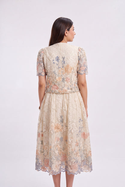 A woman viewed from the back wearing a short-sleeved midi dress with a textured floral lace pattern in beige, peach, and muted blue-gray tones, set against a plain light gray studio background.