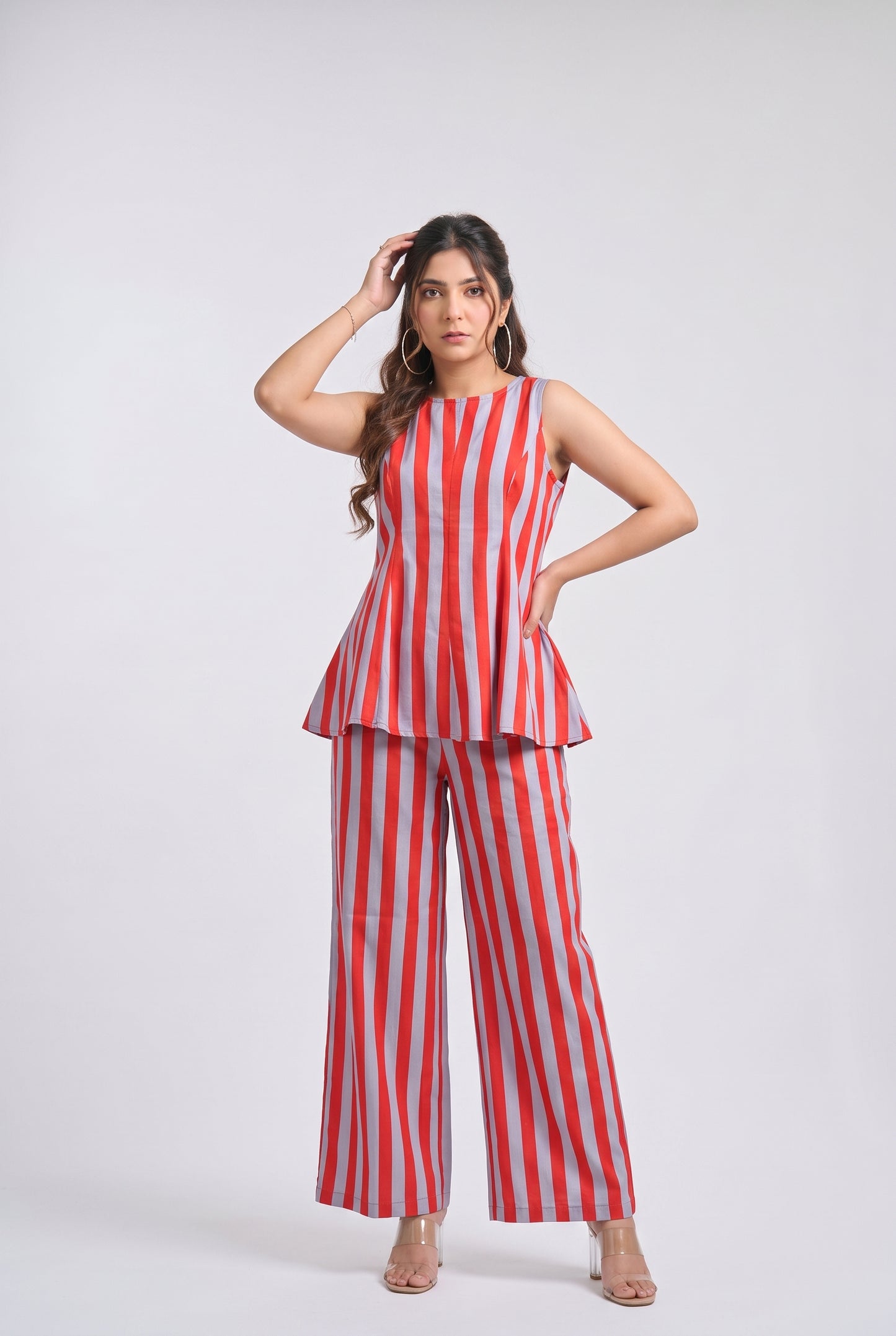 A woman modeling a sleeveless red and white vertically striped peplum top with matching wide-leg trousers, standing against a light gray studio background.