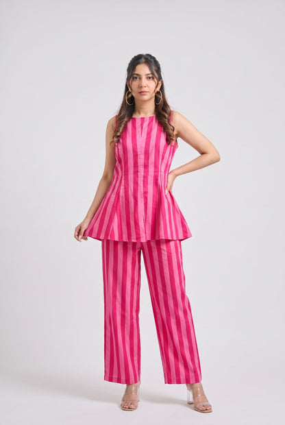 A woman modeling a pink and magenta striped sleeveless peplum top and matching wide-leg pants, styled with a clear high-heeled sandals against a light gray studio background.