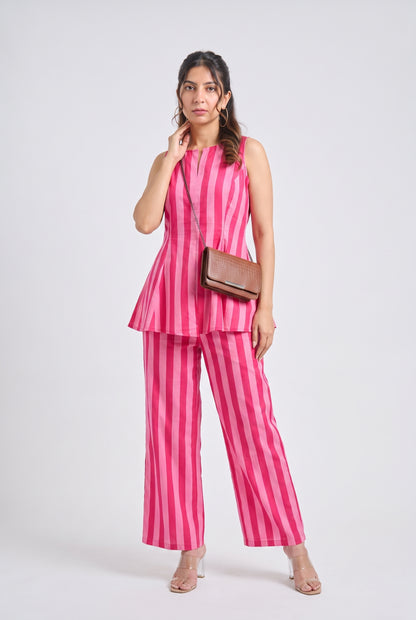 A woman modeling a pink and magenta striped sleeveless peplum top and matching wide-leg pants, styled with a brown crossbody bag and clear high-heeled sandals against a light gray studio background.