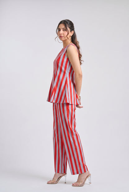 A woman modeling a sleeveless red and white vertically striped peplum top and matching wide-leg trousers, paired with clear high-heeled sandals, against a light gray studio background.