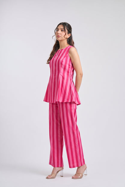 A woman modeling a sleeveless pink and magenta striped peplum top and matching wide-leg trousers, paired with clear high-heeled sandals, against a light gray studio background.