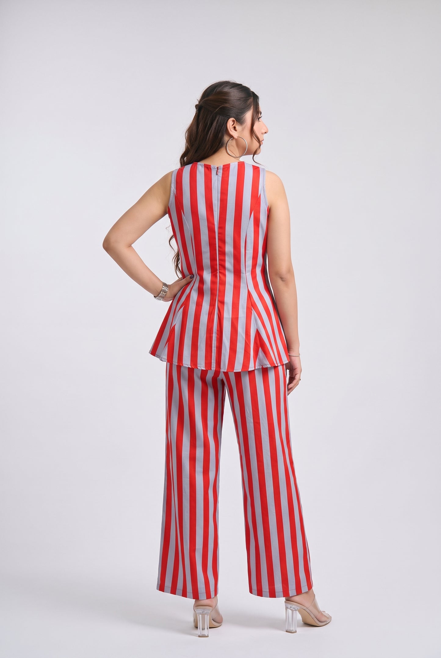 A woman is shown from the back wearing a sleeveless red and white vertically striped peplum top with matching wide-leg pants, paired with clear high-heeled sandals, against a light gray studio background.