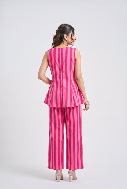 A woman is shown from the back wearing a bright pink and white vertically striped sleeveless peplum top and matching wide-leg trousers, paired with clear high-heeled sandals, against a light gray studio background.