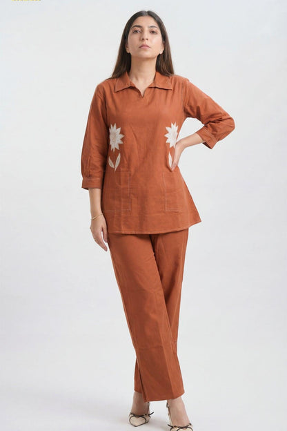 A woman modeling a rust-orange two-piece linen set consisting of a long-sleeve collared top with white floral embroidery and matching wide-leg trousers, set against a plain light gray background.