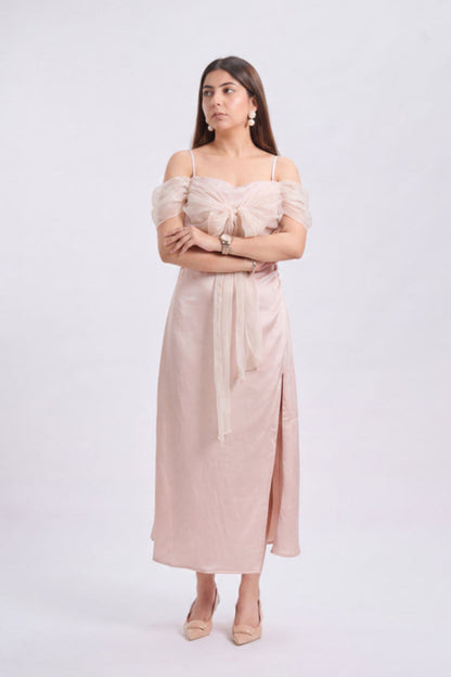 A woman modeling a light blush pink off-the-shoulder midi dress with a tie waist and side slit, standing against a plain light gray studio background.