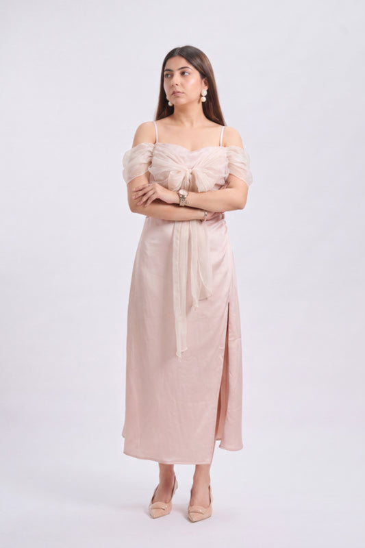 A woman modeling a light blush pink off-the-shoulder midi dress with a tie waist and side slit, standing against a plain light gray studio background.