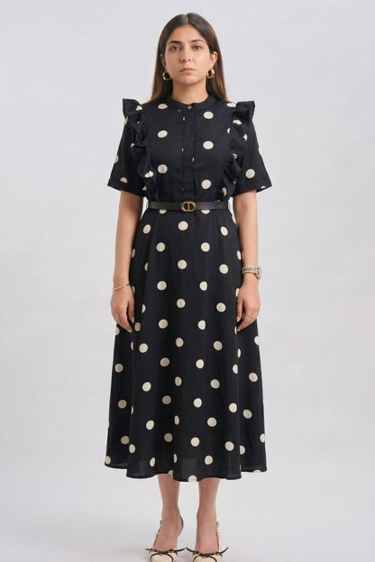 A woman modeling a black midi dress with large white polka dots, ruffled short sleeves, and a matching belt with a gold buckle, shot against a light gray studio background.