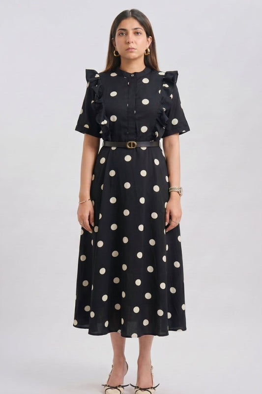 A woman modeling a black midi dress with large white polka dots, ruffled short sleeves, and a matching belt with a gold buckle, shot against a light gray studio background.