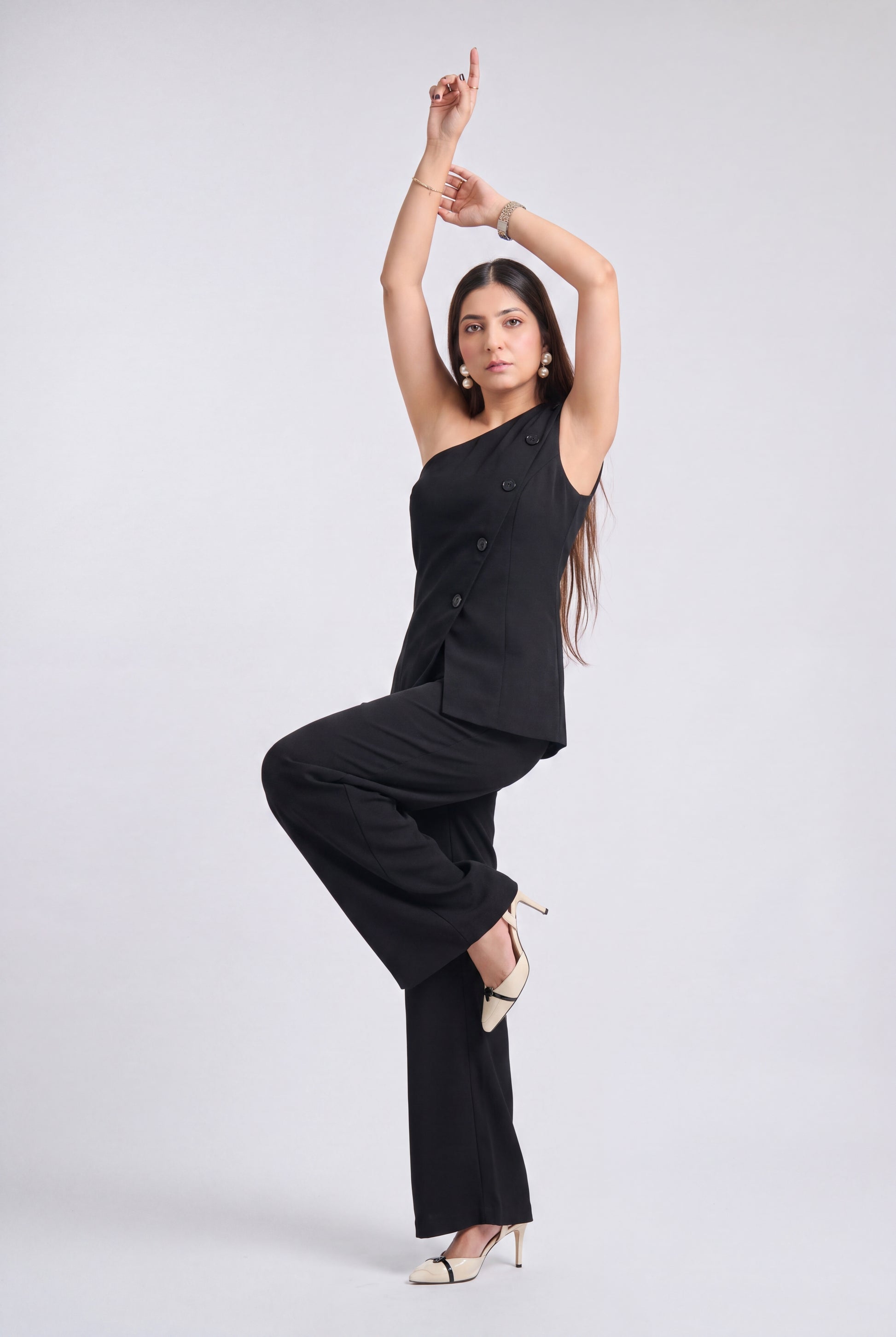 A woman modeling a black one-shoulder double-breasted blazer with matching wide-leg trousers, styled with beige high heels, against a light gray studio background.