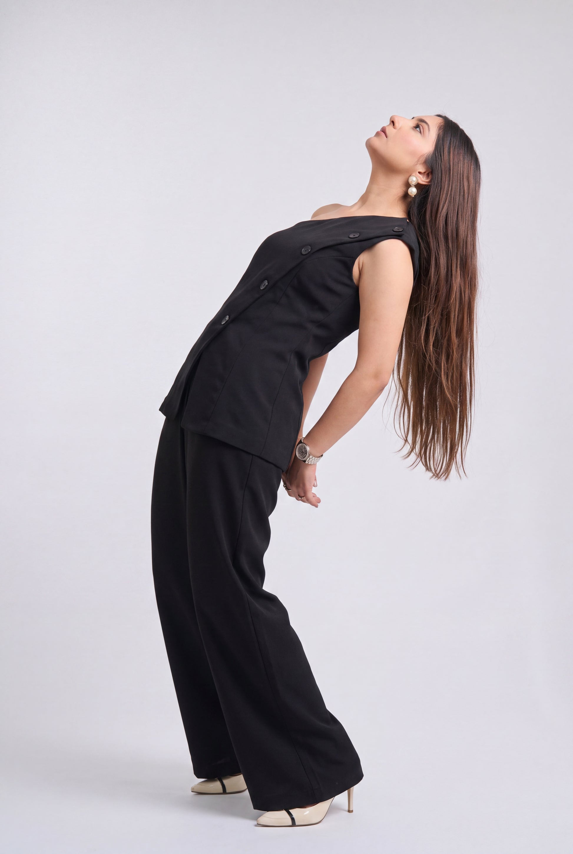 A woman modeling a black sleeveless vest and matching wide-leg trousers, posing in a studio setting against a light gray background.