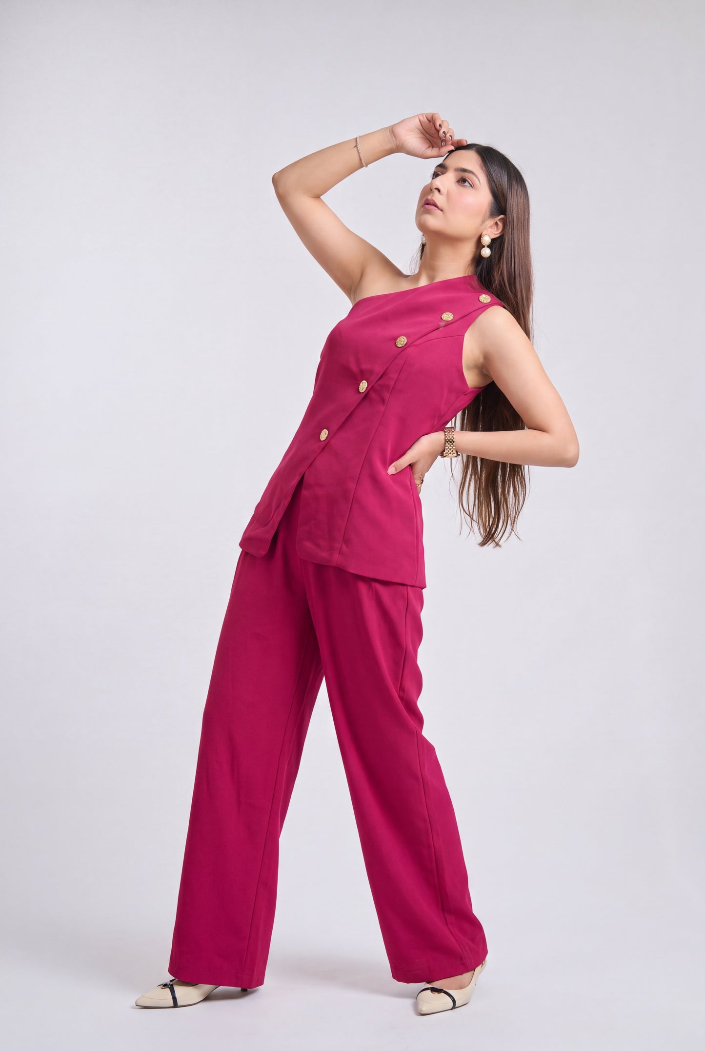 A woman modeling a bright magenta two-piece outfit consisting of a one-shoulder double-breasted top with gold buttons and matching wide-leg pants, posed against a light gray studio background.