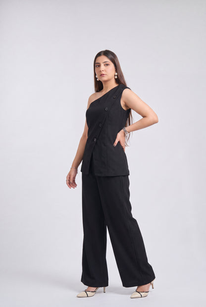 A woman modeling a black two-piece outfit consisting of a one-shoulder sleeveless top with buttons and matching wide-leg pants, photographed against a light gray studio background.