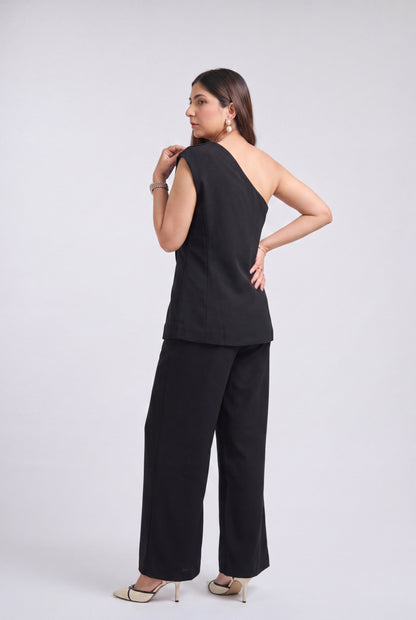 A woman modeling a black one-shoulder sleeveless top and matching wide-leg trousers, shown from a side-back angle against a light gray studio background.