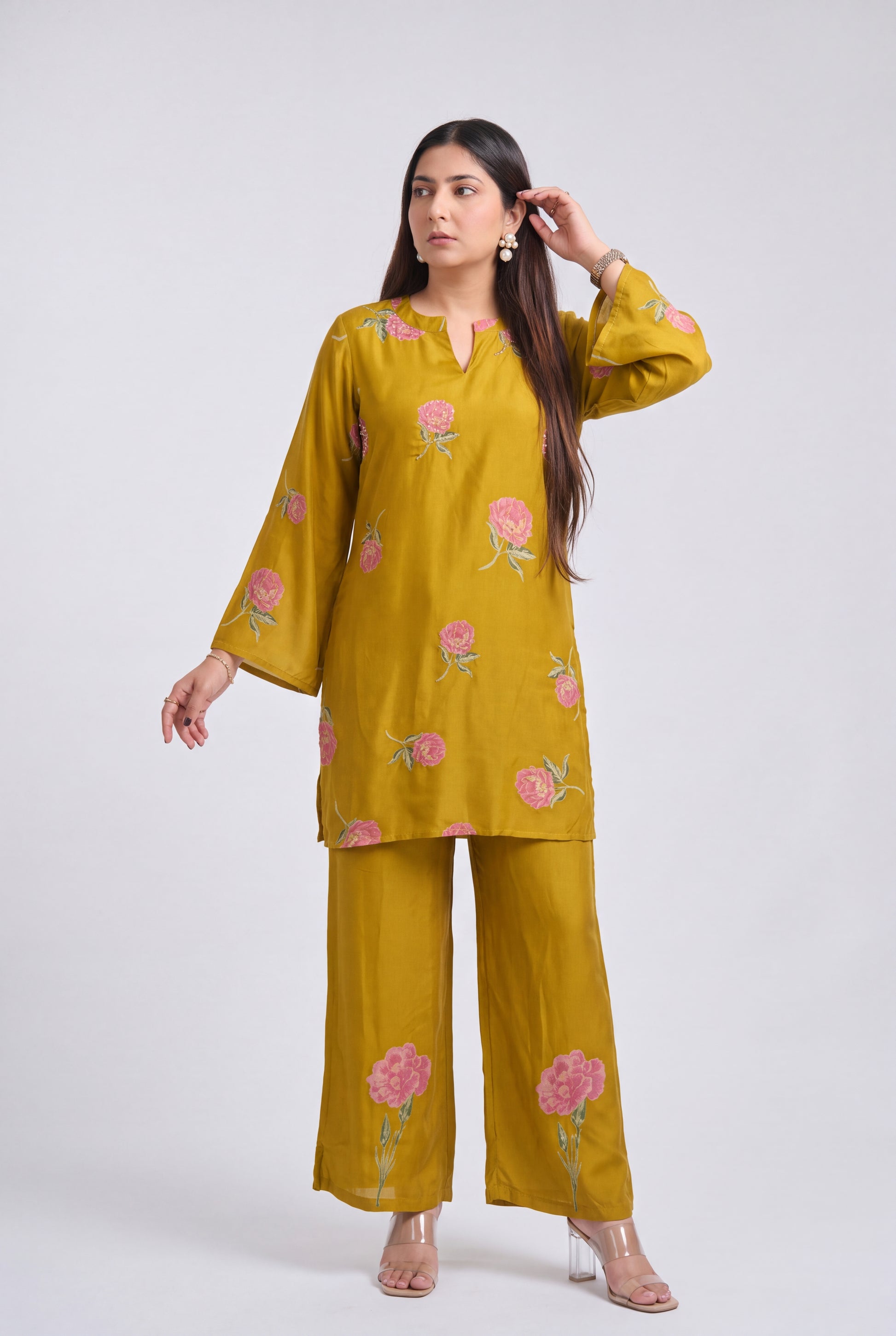 A woman modeling a mustard yellow kurta and trouser set with pink floral embroidery, set against a plain light gray studio background.