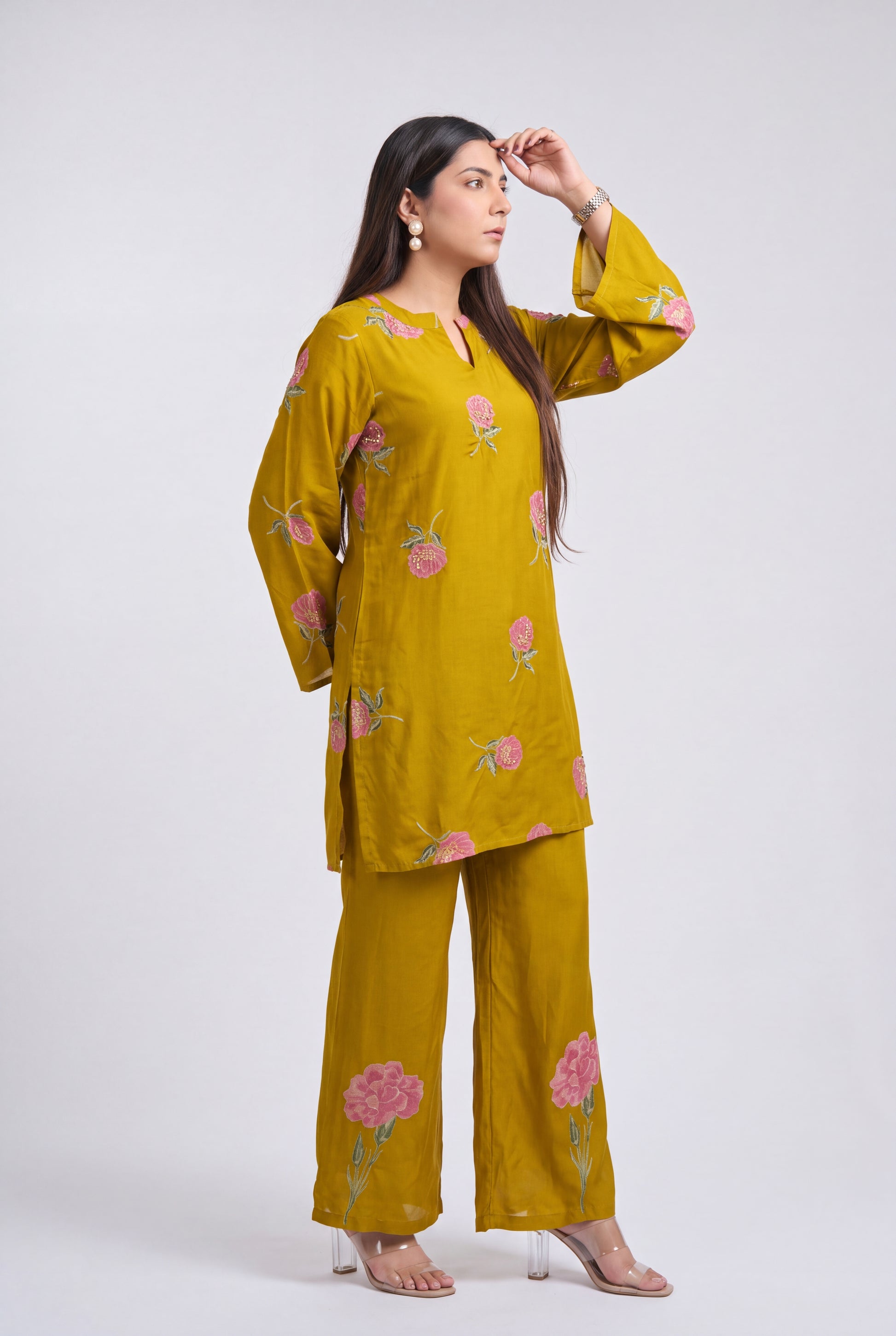 A woman modeling a mustard yellow two-piece kurta and pant set with pink floral embroidery, standing against a light gray studio background.