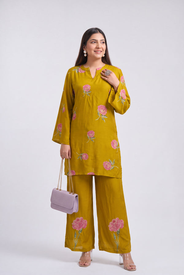 A woman modeling a mustard yellow two-piece kurta set with pink floral embroidery, holding a light purple handbag against a plain light gray studio background.