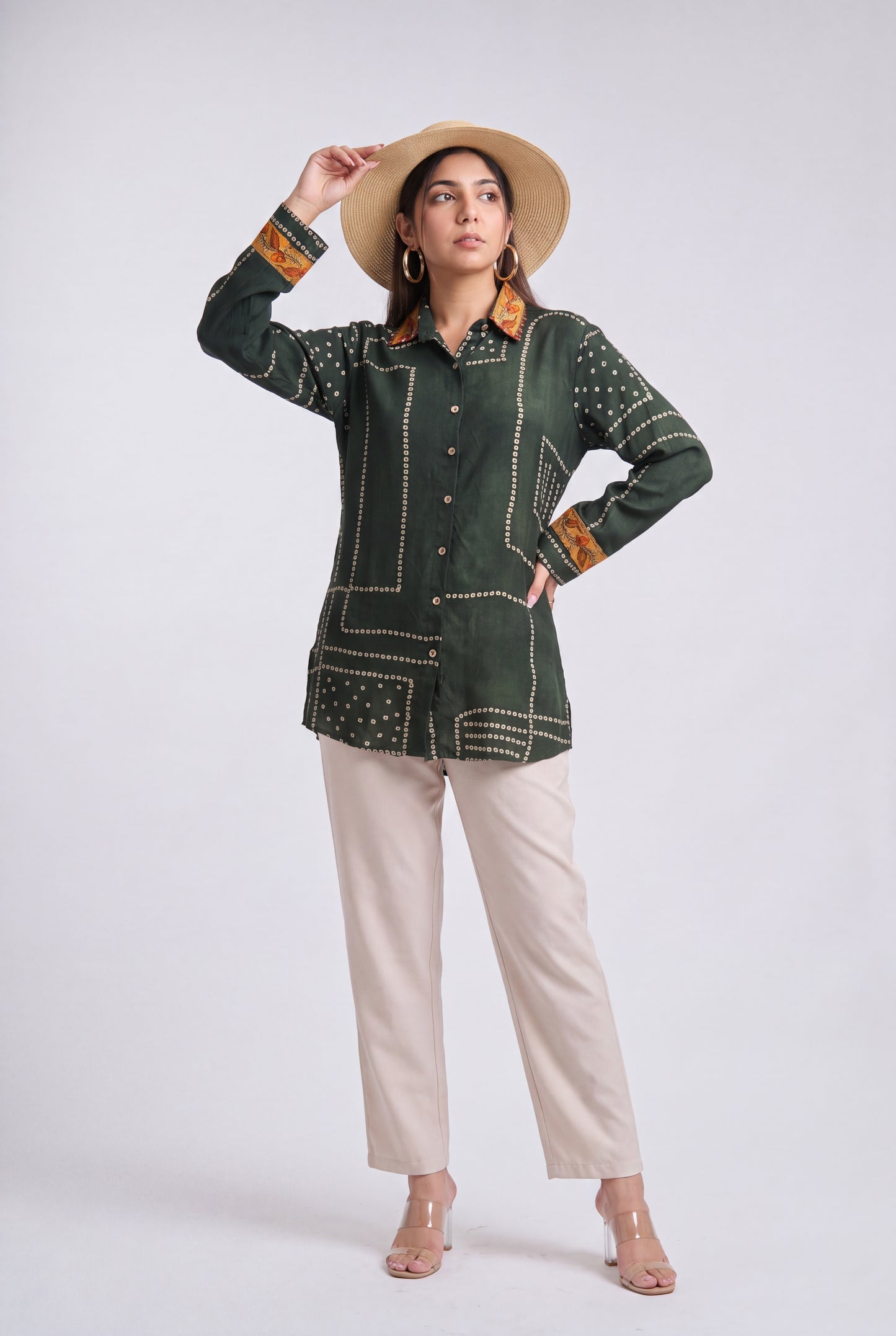 A woman modeling a dark green embroidered long-sleeve shirt with beige pants, a straw hat, and clear high-heeled sandals against a light gray studio background.