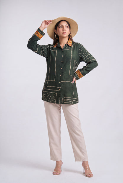 A woman modeling a dark green embroidered long-sleeve shirt with beige pants, a straw hat, and clear high-heeled sandals against a light gray studio background.