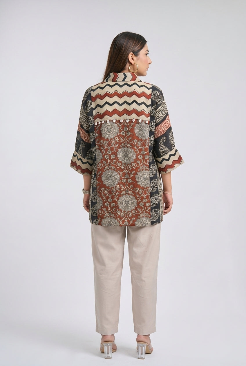 A woman viewed from the back wearing a short-sleeve button-up shirt with a complex geometric and floral patchwork print in rust red, black, beige, and white, paired with beige trousers and clear high-heeled sandals, set against a plain light gray studio background.