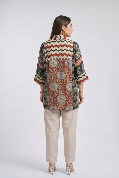 A woman viewed from the back wearing a short-sleeve button-up shirt with a complex geometric and floral patchwork print in rust red, black, beige, and white, paired with beige trousers and clear high-heeled sandals, set against a plain light gray studio background.