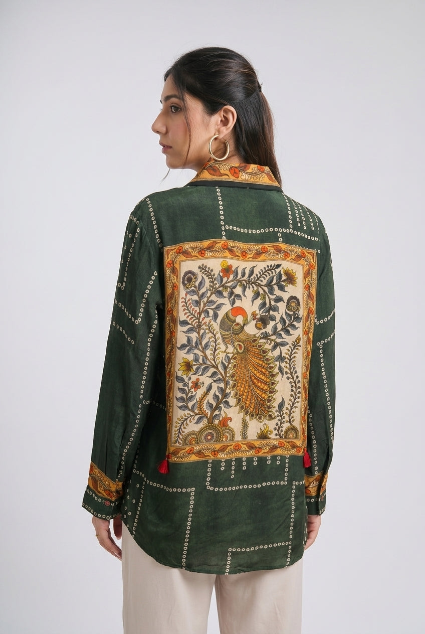 A woman shown from the back wearing a dark green embroidered jacket with a large peacock motif on the back panel, paired with light beige pants, against a plain light gray background.