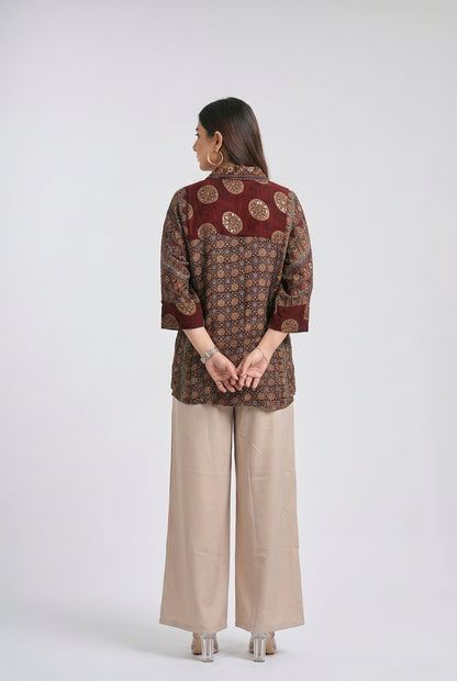 A woman is shown from the back wearing a patterned rust-red and brown printed shirt with beige wide-leg trousers, standing against a light gray studio background.
