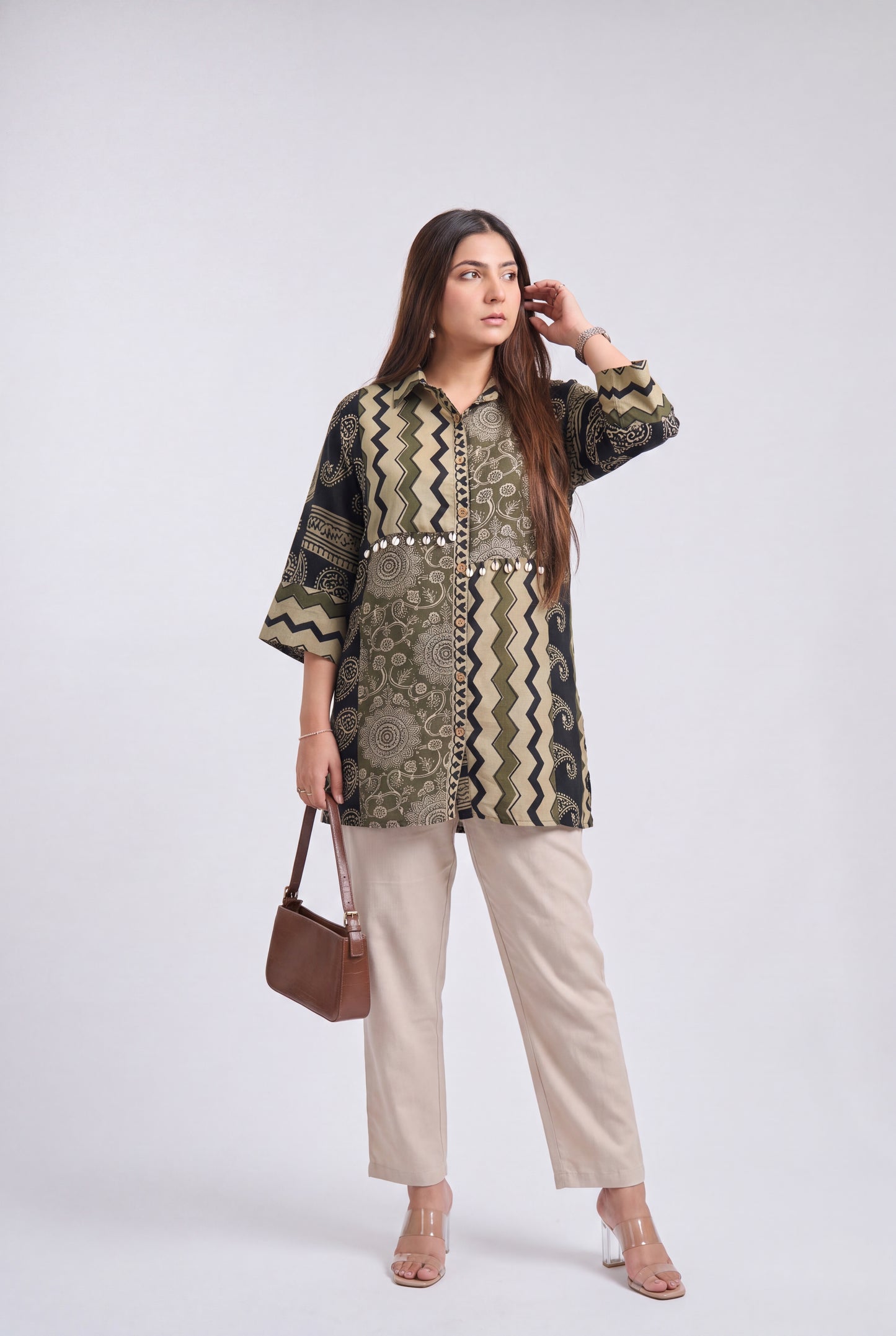 A woman models a patterned button-up tunic top with beige trousers, holding a brown handbag, against a light gray studio background.