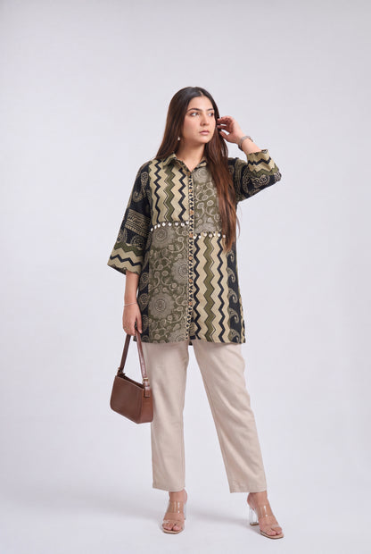 A woman models a patterned button-up tunic top with beige trousers, holding a brown handbag, against a light gray studio background.