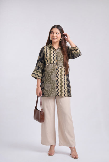 A woman modeling a patterned button-up shirt with beige wide-leg pants, holding a brown handbag against a light gray studio background.