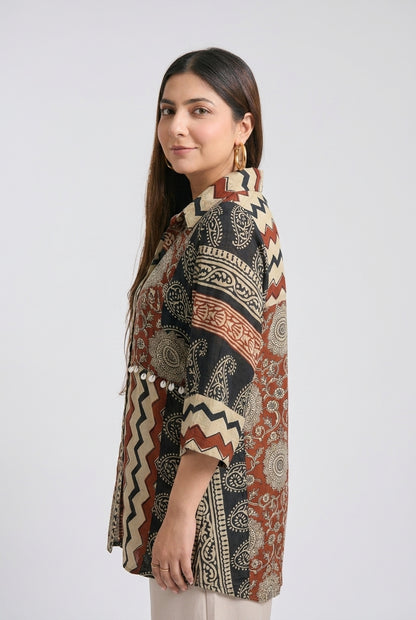 A woman modeling a short-sleeved kurta with a complex multicolor block print pattern featuring black, rust red, beige, and white geometric and paisley motifs, set against a plain light gray background.