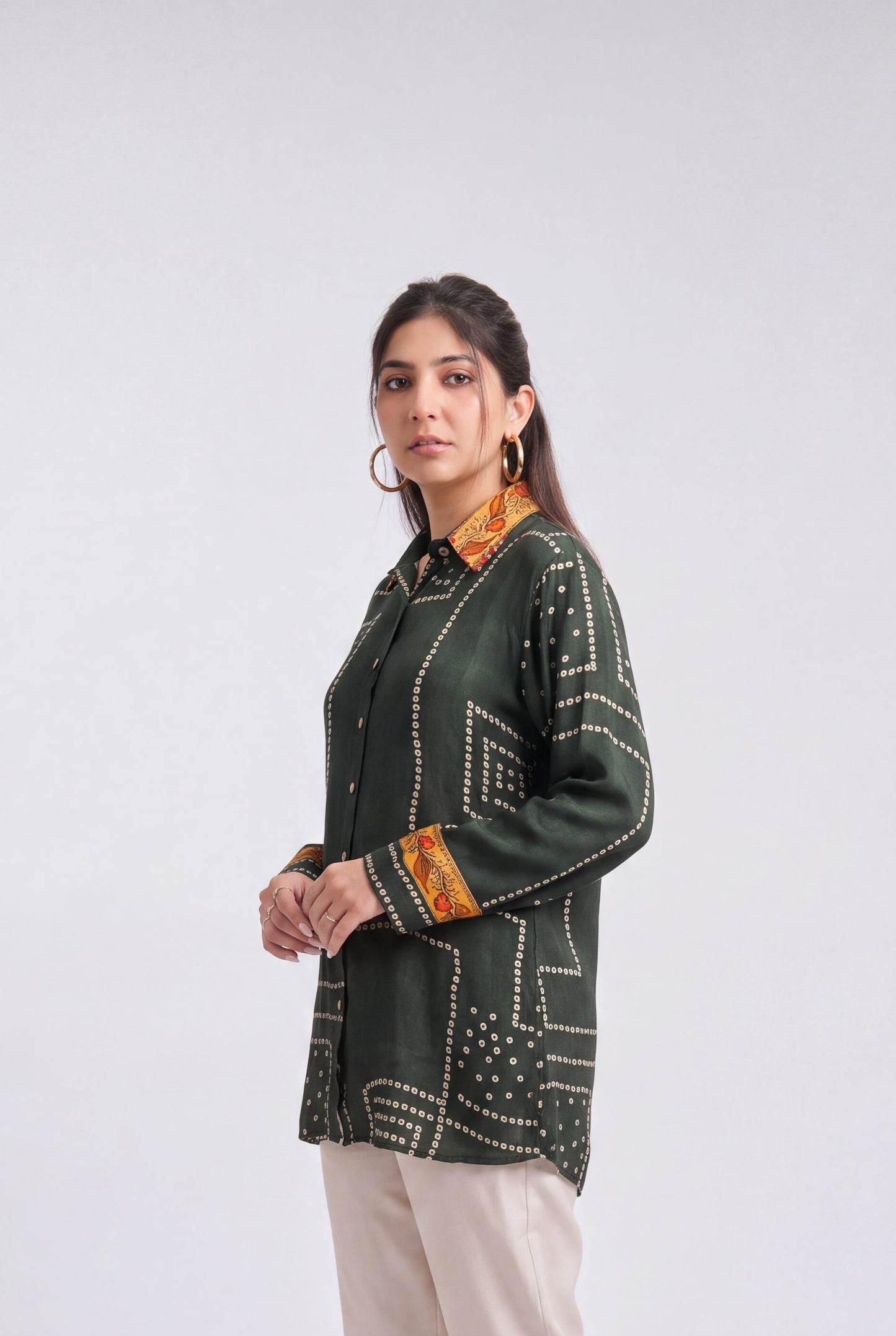 A woman modeling a dark green printed kurta with orange and white geometric patterns, paired with beige trousers and clear high-heeled sandals, against a light gray studio background.