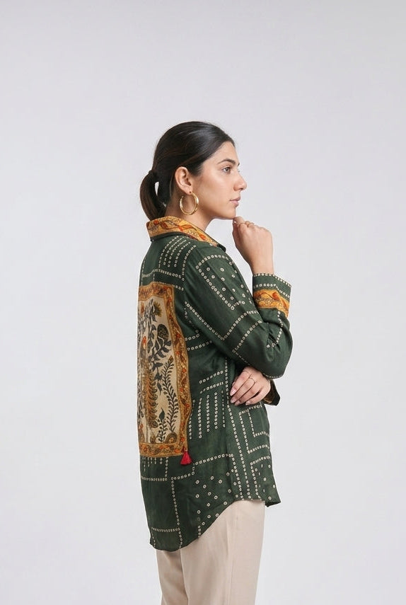 A woman stands in profile wearing a dark green patterned long-sleeve top with a large tan and gold patchwork panel on the back, paired with beige trousers and clear high-heeled sandals, against a light gray studio background.