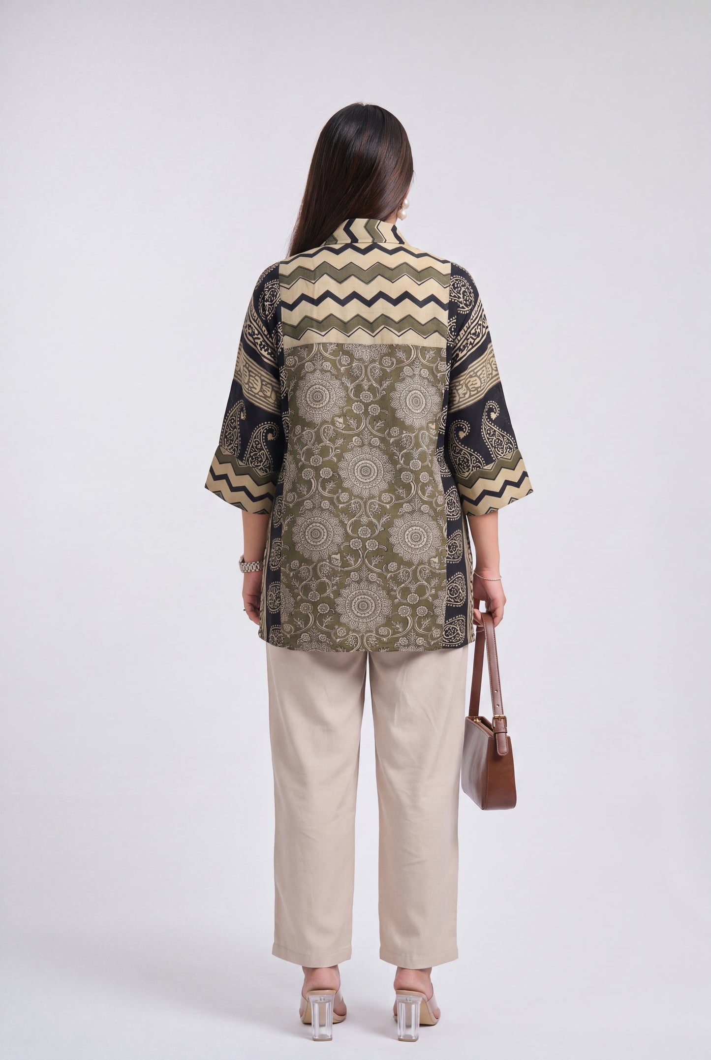 A woman shown from the back wearing a patterned short-sleeve button-up shirt with beige, olive green, and black designs, paired with beige wide-leg pants and holding a brown handbag against a light gray studio background.