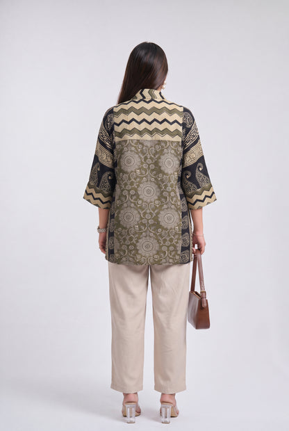 A woman shown from the back wearing a patterned short-sleeve button-up shirt with beige, olive green, and black designs, paired with beige wide-leg pants and holding a brown handbag against a light gray studio background.