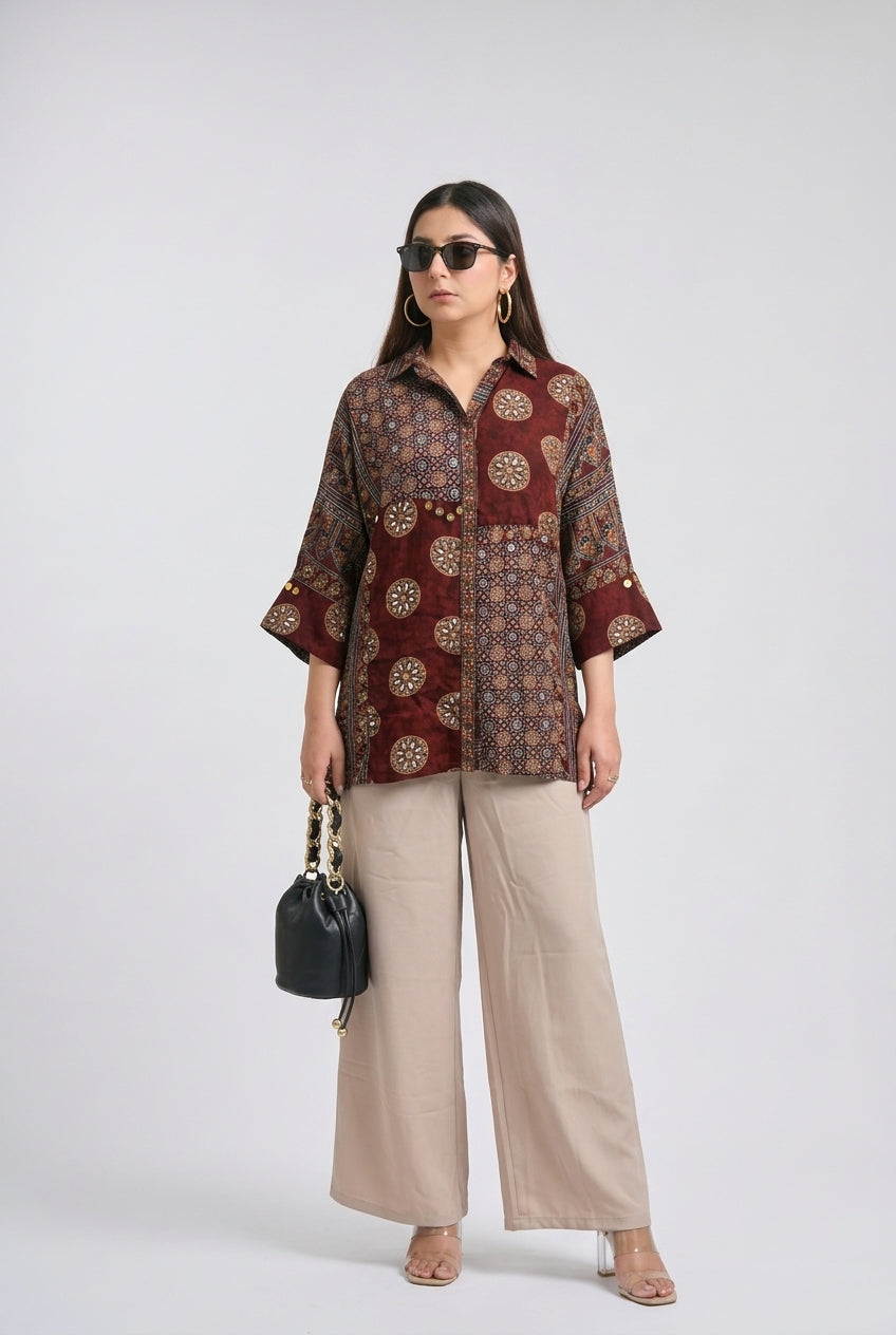 A woman modeling a patterned button-up shirt with three-quarter sleeves, paired with wide-leg beige trousers, sunglasses, and a small black handbag, against a light gray studio background.