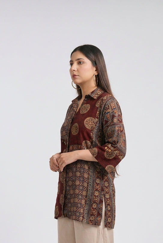 A woman modeling a three-quarter sleeve kurta with a brown and gold circular pattern over a burgundy base, paired with beige wide-leg trousers and clear high-heeled sandals, set against a plain light gray studio background.