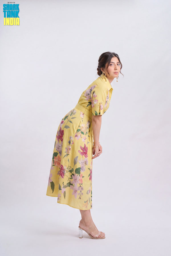 A woman modeling a yellow midi dress with a pink and green floral print, standing in a side-profile pose against a plain white studio background. A 'Think Think India' logo is visible in the top left corner.