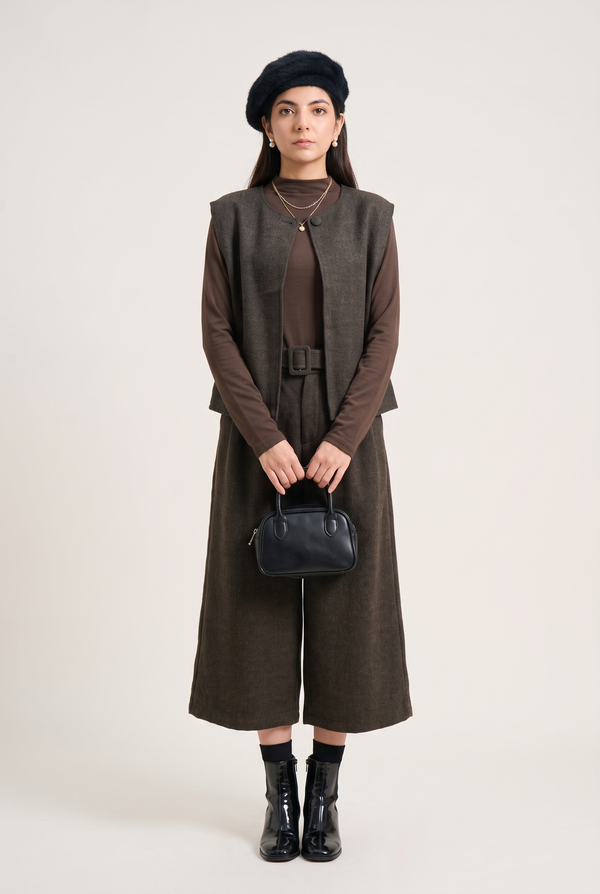 A model stands against a light beige studio background wearing a dark brown long-sleeve vest over a matching brown dress, accessorized with a black beret, a small black handbag, and black ankle boots.
