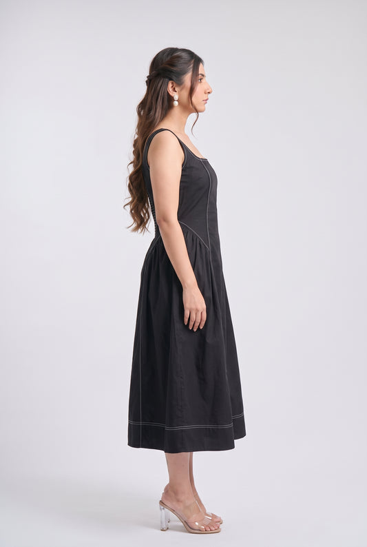 A woman modeling a sleeveless black midi dress with white contrast stitching, shown in profile against a light gray studio background.