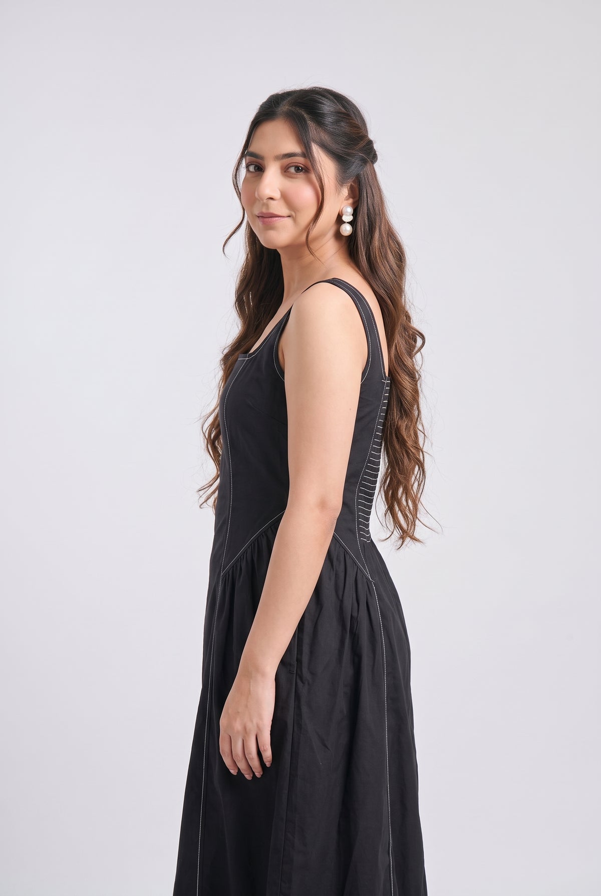 A woman modeling a sleeveless black midi dress with structured shoulder straps and a gathered skirt, photographed against a light gray studio background.