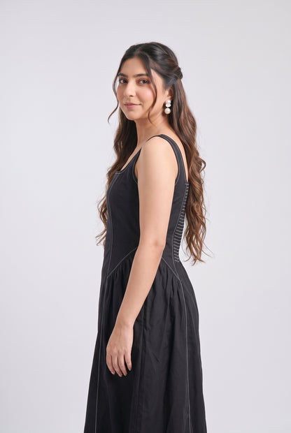 A woman modeling a sleeveless black midi dress with structured shoulder straps and a gathered skirt, photographed against a light gray studio background.