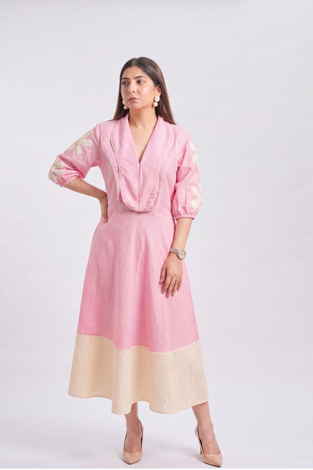 A woman modeling a pink and beige color-blocked midi dress with embroidered sleeves, standing against a light gray studio background.