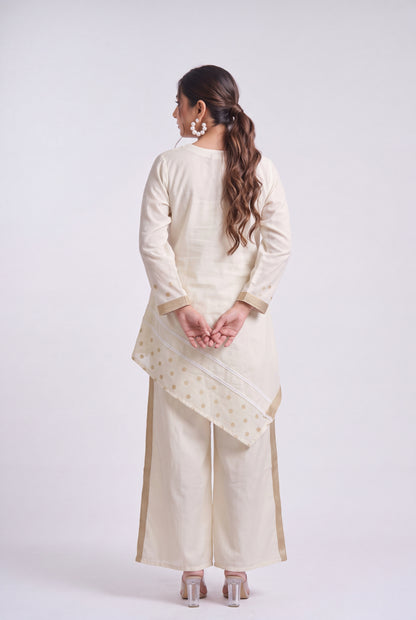 A woman is shown from the back wearing a cream-colored three-piece traditional outfit consisting of a long-sleeve kurta, wide-leg pants, and a matching dupatta with gold motifs, set against a plain light gray studio background.