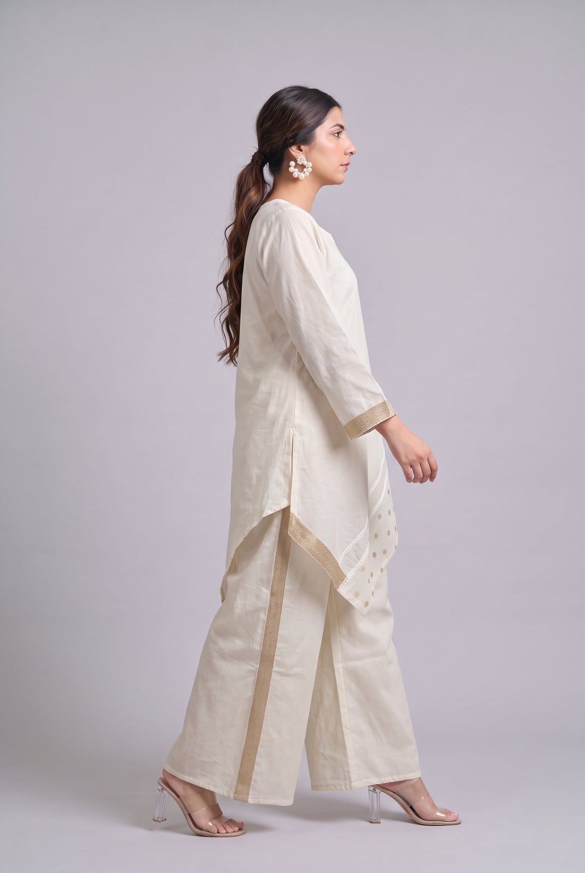 A woman modeling a cream-colored traditional outfit consisting of a long kurta, wide-leg trousers, and a matching dupatta, shown from a side profile against a light gray studio background.
