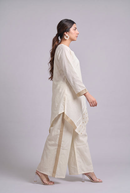 A woman modeling a cream-colored traditional outfit consisting of a long kurta, wide-leg trousers, and a matching dupatta, shown from a side profile against a light gray studio background.
