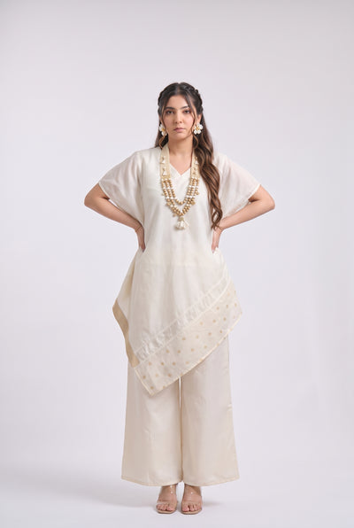 A woman modeling a cream-colored traditional outfit consisting of a short-sleeve kurta, wide-leg trousers, and a matching dupatta with gold embroidery, set against a plain light gray background.