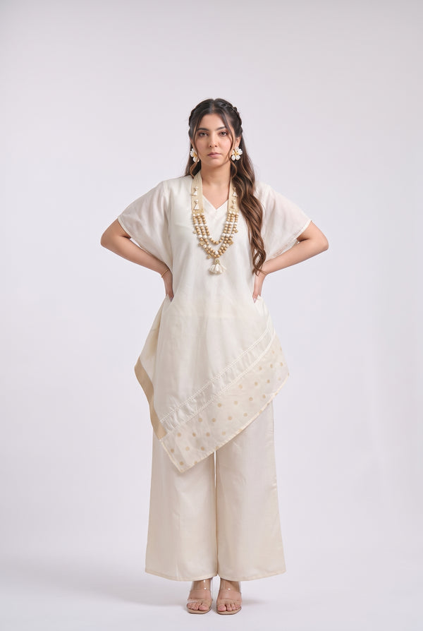A woman modeling a cream-colored traditional outfit consisting of a short-sleeve kurta, wide-leg trousers, and a matching dupatta with gold embroidery, set against a plain light gray background.