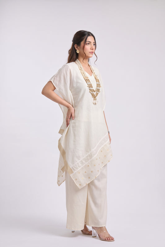 A woman modeling a cream-colored Indian ethnic outfit consisting of a kurta, wide-leg pants, and a dupatta, styled with gold jewelry against a light gray studio background.
