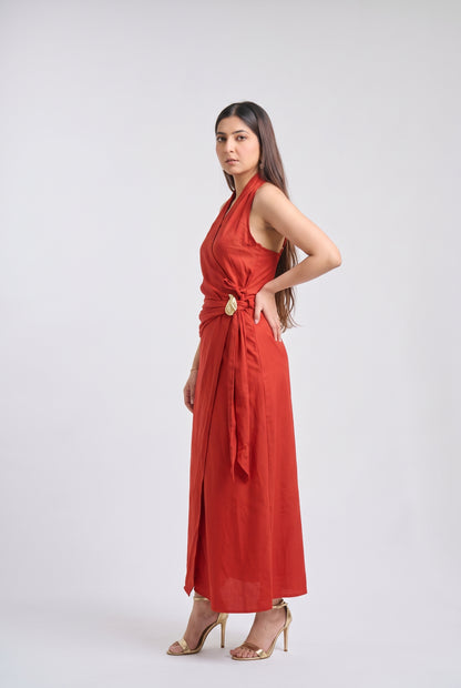 A woman modeling a sleeveless red maxi dress with a draped wrap-style waist and a large round gold accent at the center, standing against a light gray studio background.